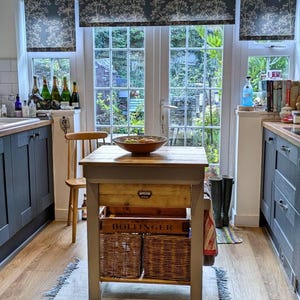 May include: A kitchen interior with a wooden table, drawer, and two wicker baskets. The table is in front of a window overlooking a garden. Dark blue cabinets and wooden flooring are visible. A wooden chair is positioned near the table. The table is 80 cm in height.
