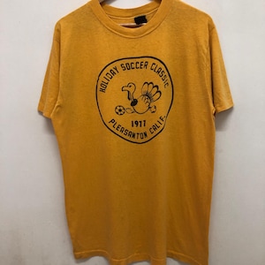 May include: A yellow t-shirt with a black circle graphic that says "Holiday Soccer Classic 1977 Pleasanton Calif." and features a cartoon turkey kicking a football.