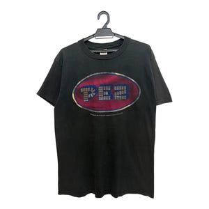 May include: A black short-sleeve t-shirt featuring a central graphic of a red oval with a metallic border. Inside the oval, the word PEZ is printed in a blocky, textured font. The shirt is displayed on a black plastic hanger against a white background.