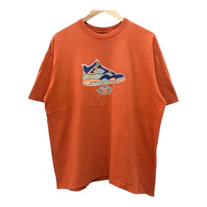 Puede incluir: Camiseta naranja con un gráfico de una zapatilla de baloncesto azul, naranja y blanca. La zapatilla está impresa en la parte delantera de la camiseta.