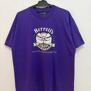 Puede incluir: Una camiseta morada con el texto "Herrell's Ice Cream" en un diseño blanco y negro. El texto "Allston, Massachusetts" está debajo del logotipo. La camiseta está colgada de una percha negra.