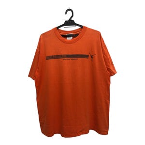 May include: Orange t-shirt with a black graphic and text. The text reads "AUSTIN ATHLETIC CLUB 2004" above a horizontal black line and a silhouette of a person. The shirt is hanging on a black hanger.