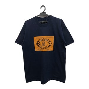Op de afbeelding: Marineblauw T-shirt met een rechthoekige oranje patch met de tekst "RUBBER SOUL" in een gestileerd lettertype. De patch bevat ook de tekst "PAST PRESENT FUTURE" en "WORLDWIDE APPAREL". Het T-shirt hangt aan een zwarte hanger.