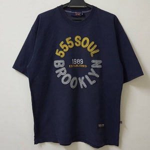 May include: Navy blue short-sleeved t-shirt with a circular design featuring "555 SOUL BROOKLYN 1989 ESTABLISHED" in yellow, white, and grey. The shirt is hanging on a black hanger.