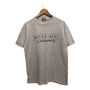 Op de afbeelding: Een grijs T-shirt met korte mouwen met de tekst "ROVER MINI" en "MINI COOPER" bedrukt op de voorkant. Het shirt heeft ook een klein logo en een Union Jack-vlag.