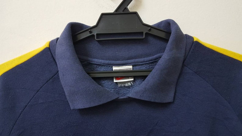 May include: A navy blue sweatshirt with yellow stripes on the sleeves. The sweatshirt has a collared neckline and a small tag on the front.