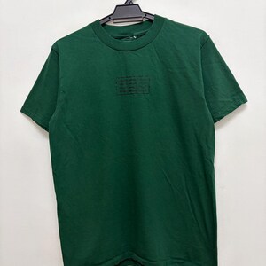 May include: A dark green t-shirt with a crew neck and short sleeves. A rectangular black graphic is centered on the chest. The shirt is hanging on a black hanger against a white background.
