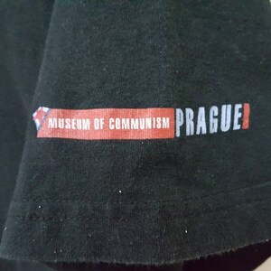 K&ouml;nnte beinhalten: Schwarzes T-Shirt mit dem Aufdruck "MUSEUM OF COMMUNISM PRAGUE" auf dem &Auml;rmel. Der Text ist in Wei&szlig; und Rot gehalten, mit einem kleinen grafischen Element. Das Shirt scheint aus Baumwolle zu sein.