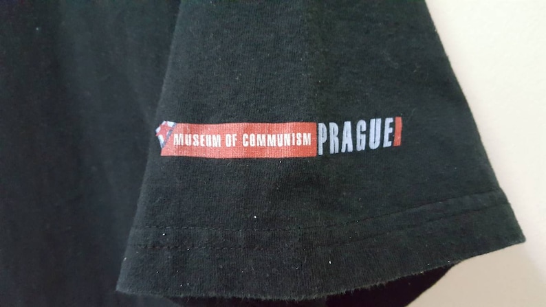 K&ouml;nnte beinhalten: Schwarzes T-Shirt mit dem Aufdruck "MUSEUM OF COMMUNISM PRAGUE" auf dem &Auml;rmel. Der Text ist in Wei&szlig; und Rot gehalten, mit einem kleinen grafischen Element. Das Shirt scheint aus Baumwolle zu sein.
