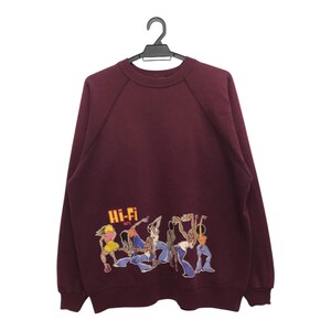 May include: A burgundy sweatshirt featuring a graphic of dancing figures with musical instruments. The text "Hi-Fi" is in yellow and orange. The sweatshirt is suspended from a black hanger.