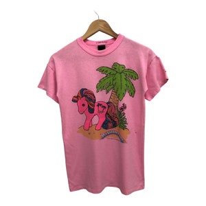 Op de afbeelding: Roze T-shirt met een afbeelding van een regenboogkleurig pony dat onder een palmboom staat. De tekst "My Little Pony" staat onder de pony gedrukt.