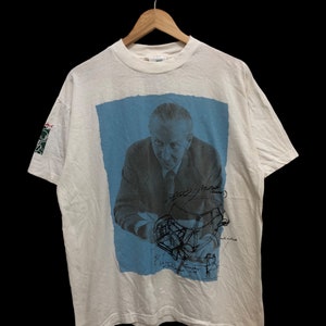 Op de afbeelding: Wit T-shirt met een blauwe en witte afbeelding van een man met een handtekening en een tekening van een auto. De tekst op het shirt luidt "Walter Gropius"