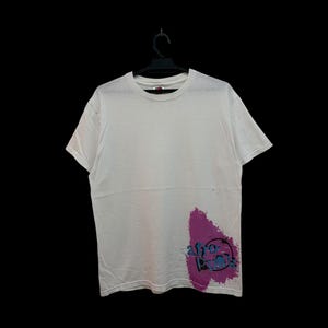 Könnte beinhalten: Weißes T-Shirt mit Rundhalsausschnitt und kurzen Ärmeln. Auf der unteren linken Seite des Shirts befindet sich ein lila-blaues Grafikdesign. Die Grafik enthält den Text "afro punk". Das Shirt hängt an einem schwarzen Kleiderbügel.