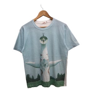 May include: A light blue t-shirt with a graphic print of a white, green, and red sculpture. The sculpture is a stylized human figure with a large head and outstretched arms.