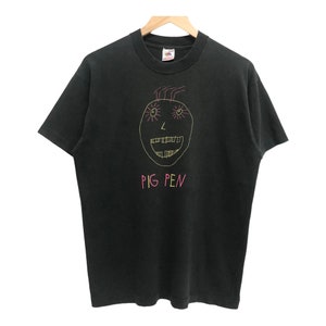 May include: Black t-shirt featuring a pink and yellow cartoon face and the text "PIG PEN" in pink.