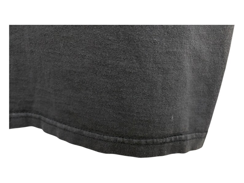 Puede incluir: Primer plano de una camiseta gris oscuro. La tela tiene un aspecto ligeramente texturizado, con costuras visibles a lo largo del dobladillo. La imagen se centra en la parte inferior de la camiseta, destacando la textura y la construcci&oacute;n.