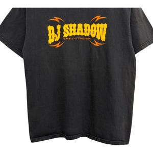 Puede incluir: Camiseta negra con el texto "DJ Shadow" en amarillo, con dise&ntilde;os de rayos naranjas. El texto "THE OUTSIDER" est&aacute; debajo del texto principal. La camiseta tiene un aspecto vintage y desgastado.