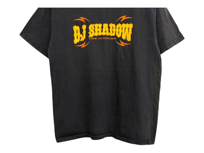 Puede incluir: Camiseta negra con el texto "DJ Shadow" en amarillo, con dise&ntilde;os de rayos naranjas. El texto "THE OUTSIDER" est&aacute; debajo del texto principal. La camiseta tiene un aspecto vintage y desgastado.
