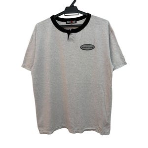 May include: A light gray short-sleeve henley shirt with a black collar and a small black button placket. The shirt features a black oval patch on the left chest with the text "EVOLUTION" printed in white, hanging on a black plastic hanger.