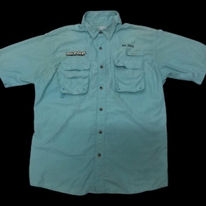 Puede incluir: Camisa de pesca azul claro de manga corta con múltiples bolsillos y cierre de botones. La camisa presenta texto bordado, incluyendo "NITRO" y "Salt Fishing". Tiene un cuello clásico y está diseñada para actividades al aire libre.