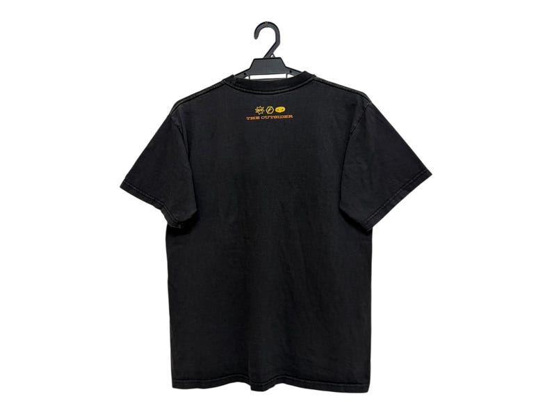 Puede incluir: Camiseta negra colgada de una percha negra. La camiseta tiene un peque&ntilde;o dise&ntilde;o amarillo y naranja en la parte superior de la espalda, con el texto "THE OUTSIDER". La camiseta es de un material suave.