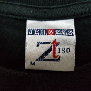 K&ouml;nnte beinhalten: Nahaufnahme eines schwarzen T-Shirt-Kragens mit einem wei&szlig;en Etikett. Das Etikett zeigt den Markennamen "JERZEES" in Blau und Rot sowie das Logo "Zt 180" in einer stilisierten Schriftart. Der Buchstabe "M" befindet sich unten.