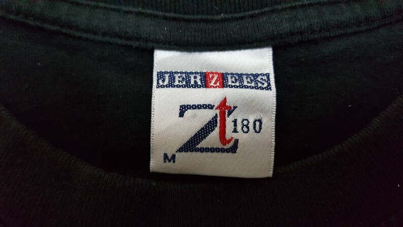 K&ouml;nnte beinhalten: Nahaufnahme eines schwarzen T-Shirt-Kragens mit einem wei&szlig;en Etikett. Das Etikett zeigt den Markennamen "JERZEES" in Blau und Rot sowie das Logo "Zt 180" in einer stilisierten Schriftart. Der Buchstabe "M" befindet sich unten.