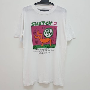 May include: White t-shirt with a graphic print of a person breakdancing with a Swatch watch on their head. The text "Swatch+ World Breakdance Championship at the Roxy September 20" is printed below the graphic.