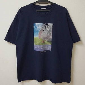 Könnte beinhalten: Marineblaues T-Shirt mit einer Grafik von Eeyore aus Winnie the Pooh. Das Bild zeigt Eeyore, der einen Marienkäfer betrachtet. Der Text "Eeyore" befindet sich unter dem Bild, darunter "Disney". Das T-Shirt hängt an einem schwarzen Kleiderbügel.