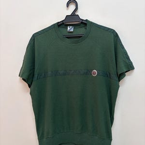May include: Dark green short-sleeved shirt with a textured knit pattern. The shirt has a dark band across the chest and a small emblem on the right. The shirt is hanging on a black hanger.