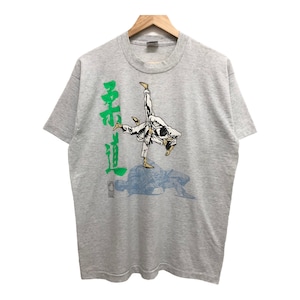 Op de afbeelding: Een grijs T-shirt met een afbeelding van een persoon in een wit martial arts uniform dat een beweging uitvoert. De tekst "柔道" staat in het groen aan de linkerkant van de afbeelding.