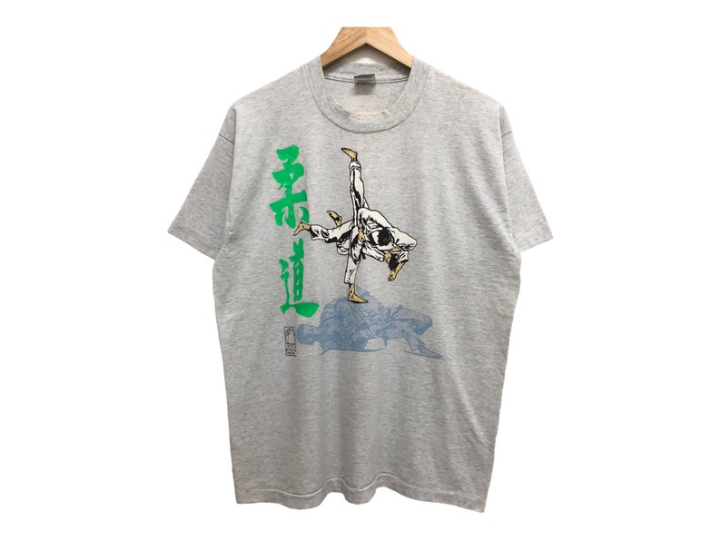 Op de afbeelding: Een grijs T-shirt met een afbeelding van een persoon in een wit martial arts uniform dat een beweging uitvoert. De tekst "柔道" staat in het groen aan de linkerkant van de afbeelding.