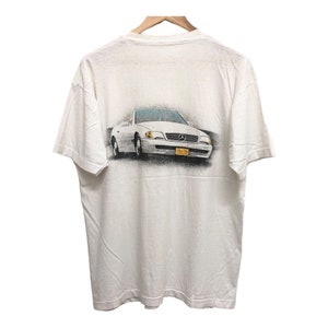 Op de afbeelding: Wit T-shirt met een grafische print van een witte Mercedes-Benz cabriolet op de achterkant. De auto heeft een geel kenteken met de tekst "Real Fun."
