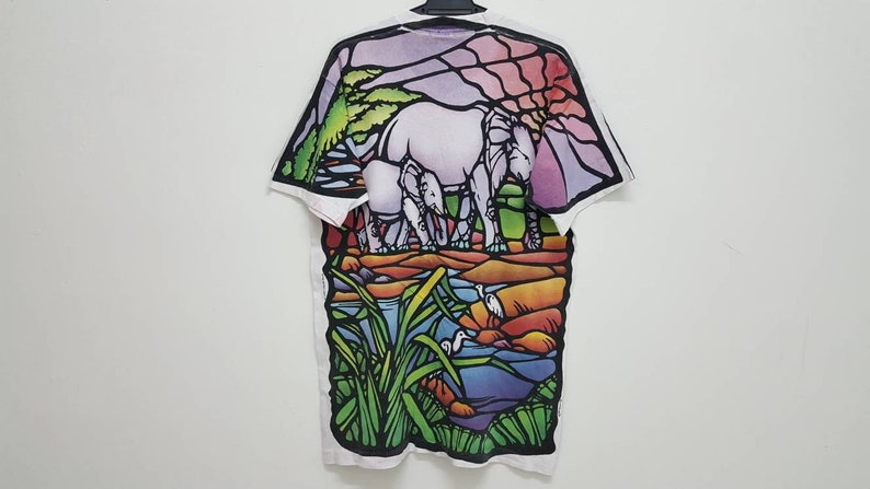 Puede incluir: Camiseta blanca con un estampado de estilo vitral de elefantes y una escena de r&iacute;o. El estampado presenta una variedad de colores, incluyendo verde, azul, morado, naranja y amarillo.