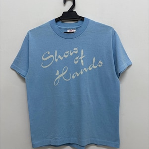 Può includere: T-shirt azzurra con la scritta "Show of Hands" in corsivo. La maglietta è appesa a una gruccia nera contro un muro bianco. La t-shirt ha maniche corte e girocollo.