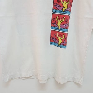 K&ouml;nnte beinhalten: Wei&szlig;es T-Shirt mit einem bunten Grafikdruck einer Person, die auf einem Delfin reitet. Die Grafik wird dreimal auf dem Shirt wiederholt.