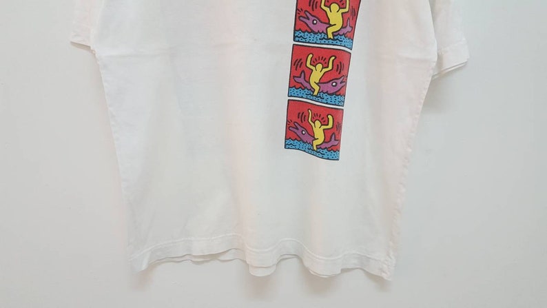 K&ouml;nnte beinhalten: Wei&szlig;es T-Shirt mit einem bunten Grafikdruck einer Person, die auf einem Delfin reitet. Die Grafik wird dreimal auf dem Shirt wiederholt.