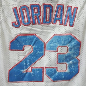 May include: White basketball jersey with blue and red trim. The back of the jersey has the name "JORDAN" and the number "23" in red with a blue and white distressed design.