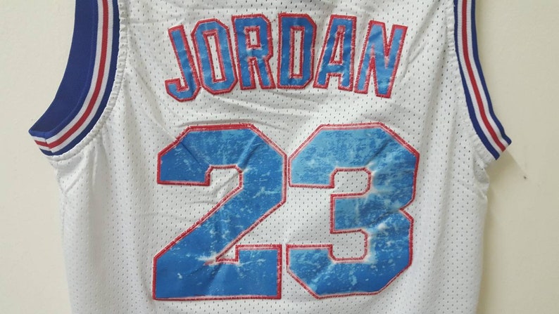 May include: White basketball jersey with blue and red trim. The back of the jersey has the name "JORDAN" and the number "23" in red with a blue and white distressed design.