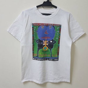 Op de afbeelding: Wit T-shirt met een kleurrijke grafische print van een cartoonfiguur dat saxofoon speelt. De tekst "NEW ORLEANS JAZZ LEGEND" en "LOUIS" zijn op de print gedrukt. De tekst "NEW ORLEANS, LOUISIANA" is onder de print gedrukt.
