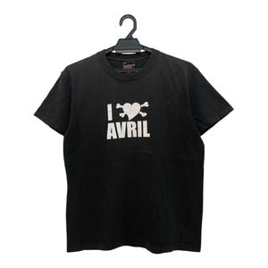 以下が含まれることがあります： 黒いTシャツ。白い文字で「I <3 AVRIL」と書かれています。ハートのマークは、2本の骨が交差したデザインと組み合わされています。Tシャツは黒いハンガーにかかっています。