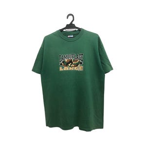 May include: A green t-shirt featuring the word "CAMOUFLAGE" above a camouflage design and the word "LARGE" below. The shirt is suspended on a black hanger against a white backdrop.