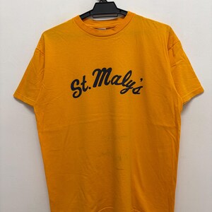 May include: A bright yellow short-sleeved t-shirt featuring the text St. Maly's printed in a dark navy blue cursive font across the chest. The garment has a crew neckline and is displayed on a black plastic hanger against a plain white wall.