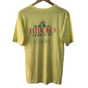Può includere: Maglietta gialla a maniche corte con una stampa grafica verde, rossa e bianca che dice "水への冒険をこえて HIROKO MORIGUCHI" e presenta un gruppo di personaggi dei cartoni animati.