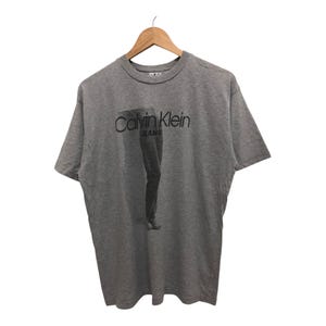 Puede incluir: Una camiseta gris de manga corta con el texto "Calvin Klein Jeans" impreso en la parte delantera. La camiseta presenta un gráfico de las piernas de una persona.