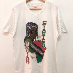 Puede incluir: Camiseta blanca con un estampado gráfico de una mujer negra con trenzas azules y blancas, llevando una diadema roja, blanca y azul. El gráfico también incluye un contorno verde, rojo y negro del continente africano con el texto "Sister of the Planet Earth" y "Nubian" escrito verticalmente.