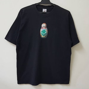 K&ouml;nnte beinhalten: Schwarzes Kurzarm-T-Shirt mit einem farbenfrohen Aufdruck einer russischen Matrjoschka-Puppe. Die Puppe hat einen &uuml;berraschten Gesichtsausdruck und ist mit gr&uuml;nen, roten und goldenen Details verziert. Das Shirt h&auml;ngt an einem schwarzen Kleiderb&uuml;gel.