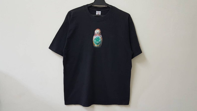 K&ouml;nnte beinhalten: Schwarzes Kurzarm-T-Shirt mit einem farbenfrohen Aufdruck einer russischen Matrjoschka-Puppe. Die Puppe hat einen &uuml;berraschten Gesichtsausdruck und ist mit gr&uuml;nen, roten und goldenen Details verziert. Das Shirt h&auml;ngt an einem schwarzen Kleiderb&uuml;gel.