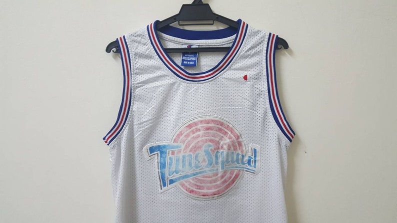 May include: White basketball jersey with red and blue trim. The jersey has a large graphic of the "Tune Squad" logo with a basketball and the words "Tune Squad" in a blue and white font.
