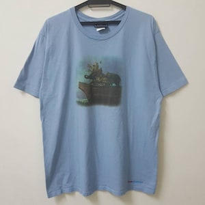 Puede incluir: Una camiseta azul claro con un estampado gráfico de un grupo de personas montando un elefante. El elefante está de pie en una plataforma con un cielo azul y nubes en el fondo.